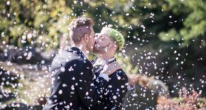 two light-skinned men kissing at their wedding with white confetti in the air