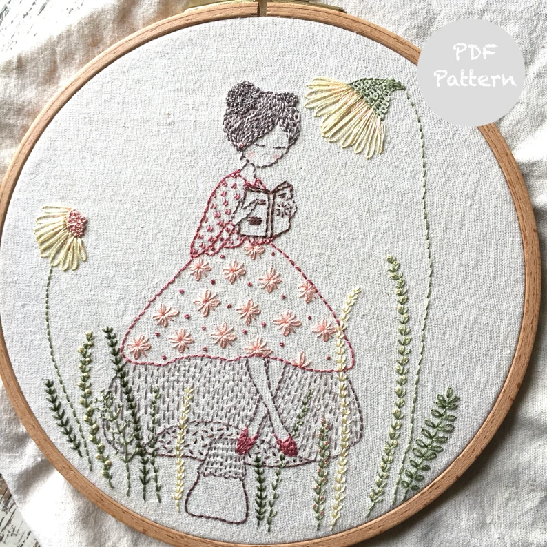 Image of an embroidery pattern featuring a person with space buns reading on a mushroom. 