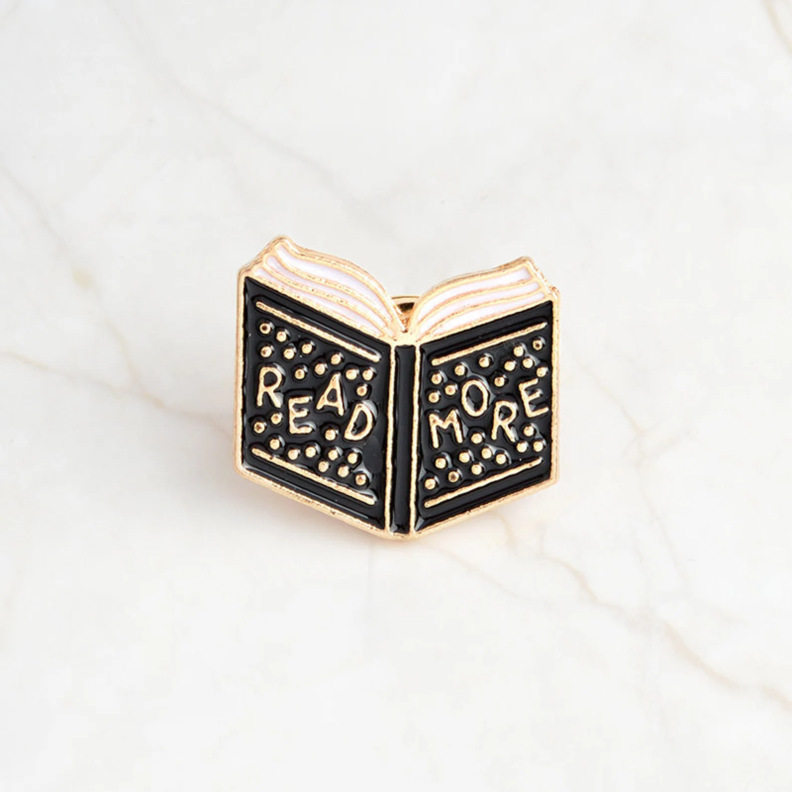 A black enamel pin in the shape of an open book that reads "Read more"