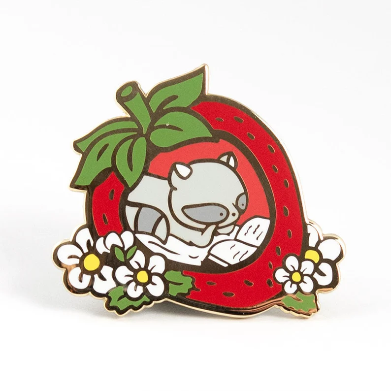 Image of a strawberry enamel pin. Inside the hollowed berry is a raccoon reading a book. 