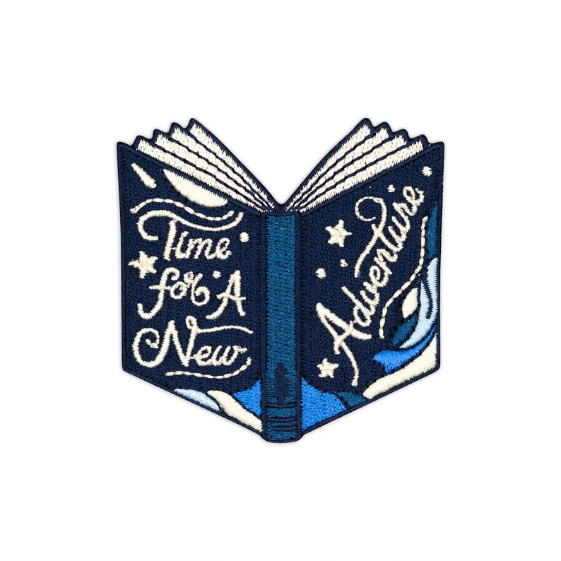 Image of a book patch in the shape of a book. It is blue and in whitefont reads "times for a new adventure."