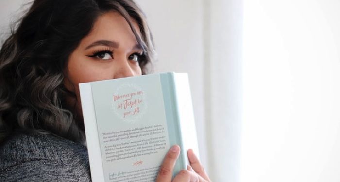 a woman with lightly-tanned skin and black eyeliner is holding an open book over the bottom half of her face and looking past the camera