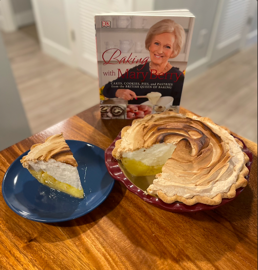 A toasty lemon meringue pie with a clean slice placed beside it on a blue plate. It set on a wooden table in front of a copy of the cookbook Baking with Mary Berry.