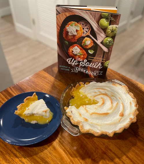 A soupy looking lemon meringue pie sits on a wooden table next to a slice on a blue plate. They sit in front of the cookbook The Up South Cookbook.