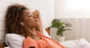 a tan-skinned Black woman relaxes in bed with her eyes closed and headphones in her ears