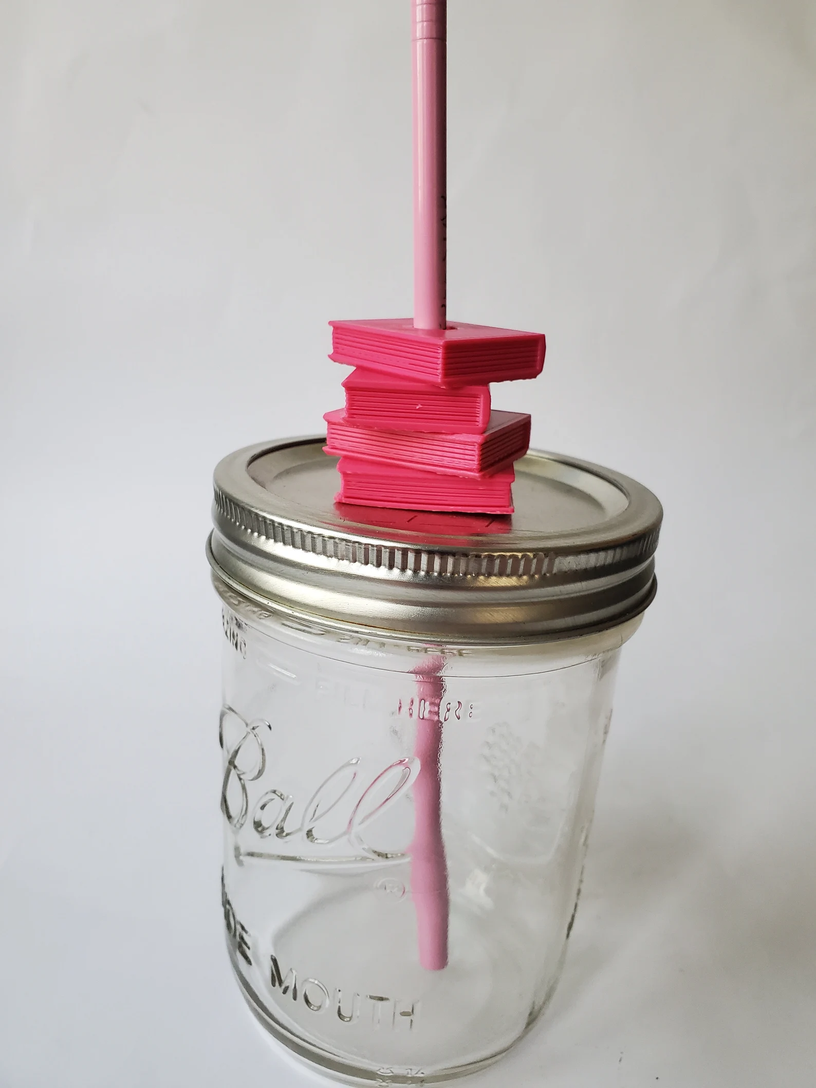 A mason jar cup with a lid, pink straw, and a straw topper in the shape of a stack of books.