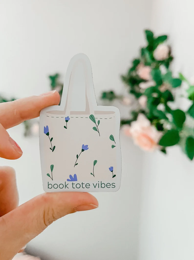 Image of a white hand holding a magnet in the shape of a tote bag. The tote reads "book tote vibes" and has purple flowers on it. 