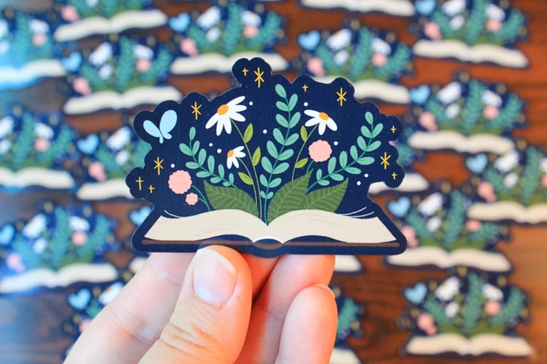 Image of a white hand holding a magnet. The magnet is in the shape of an open book, and there are flowers growing from inside the book. 