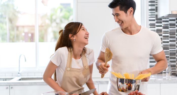 an Asian couple smiling and cooking pasta in a white kitchen