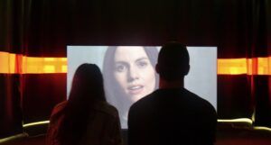 two people in a dark room watching a video of a woman speaking