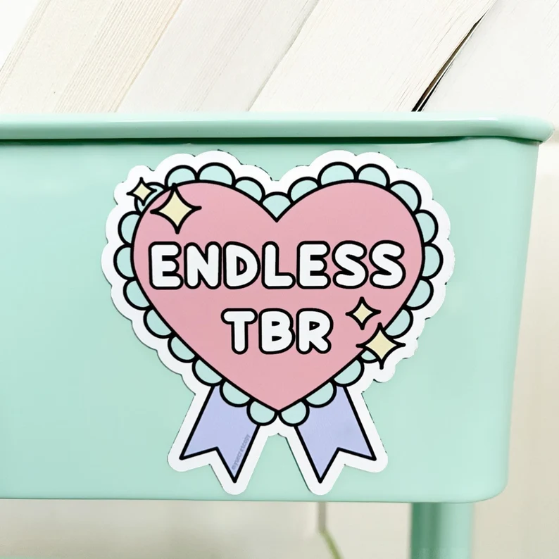 Image of a merit-badge shaped magnet. It reads "endless TBR" in the center, and it is shades of pastel pink, purple, green, and yellow. It is on a green book cart. 