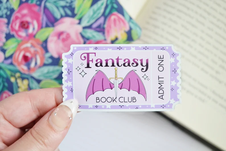 Image of a whit hand holding a magnet in the shape of a ticket. It says "fantasy book club: admit one." It features a knife and vampire-style wings. 