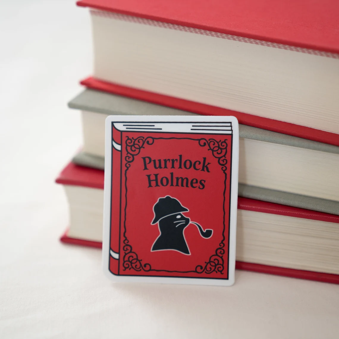photo of a red book-shaped sticker with a cat in sillhouette. above the cat is the text "Purrlock Holmes"