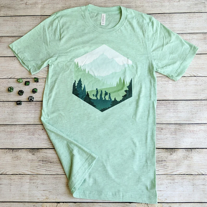 photo of a mint green tee shirt on a table next to a group of green dice. illustrated on the shirt is a group of adventurers in sillhouette trekking through a mountainous forest