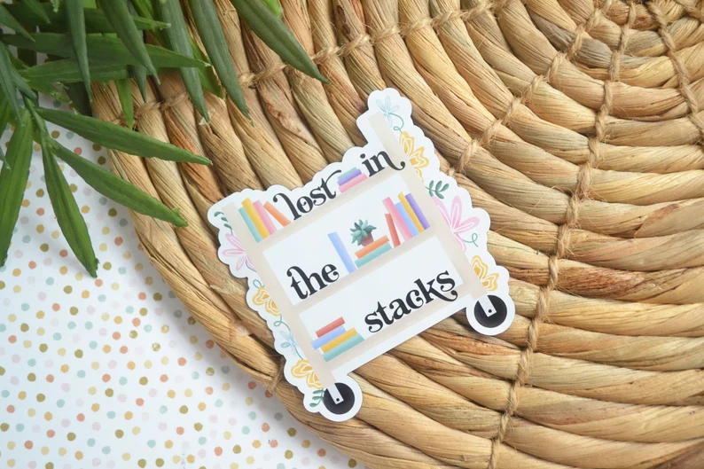 Image of a magnet in the shape of a book cart. It has flowers on the edges and on the shelves of the cart, it has books with the words "lost in the stacks."