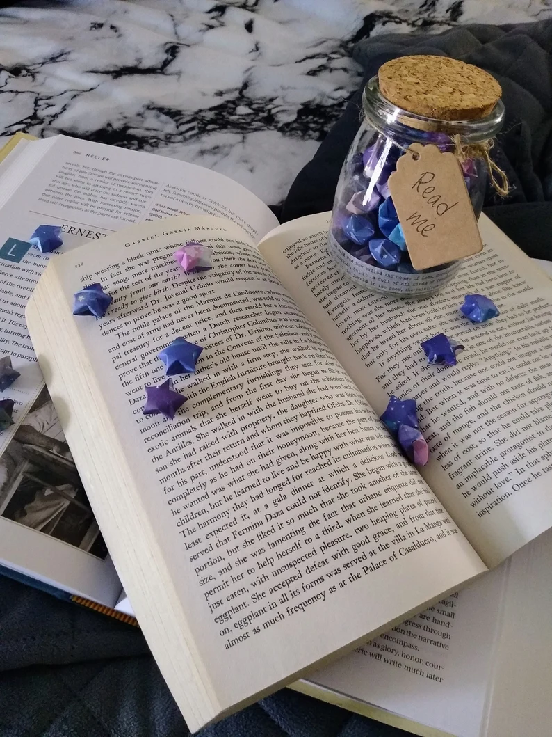 Image of a glass jar with colorful paper stars inside. The jar is on an open book and has a tag that says "read me."