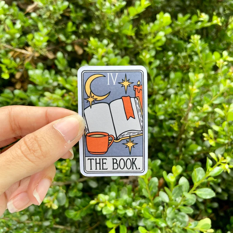 Image of a magnet designed like a tarot card. It says "the book." It's being held by a white hand in front of some greenery. 