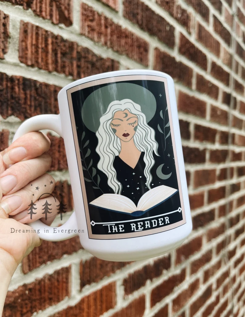 Image of a white hand holding a big white mug. The image on the mug is modeled after a tarot card called "the reader." 