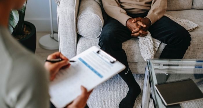 person with brown skin seen from the waist down, seated on a couch across from a person with white skin taking notes on a clipboard