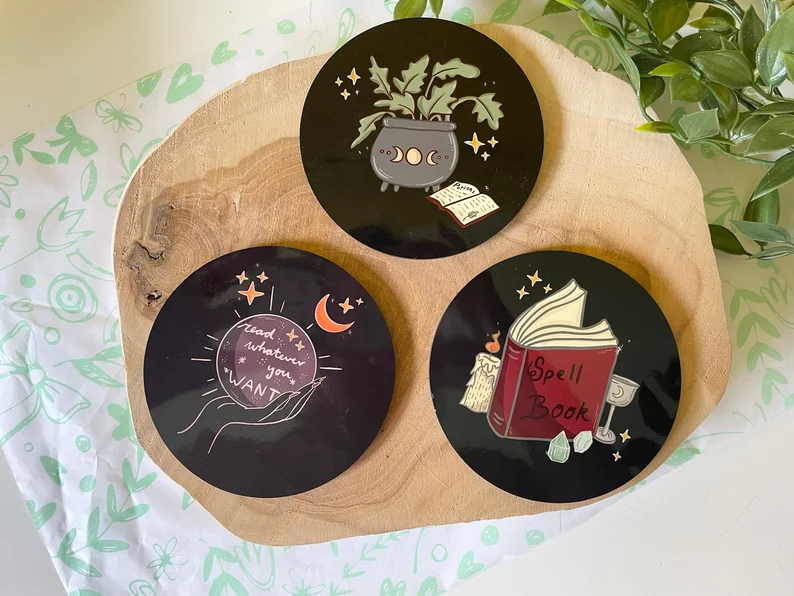 Image of three black coasters. Each has witchy images, including a magic 8 ball, a spell book, and a cauldron. 
