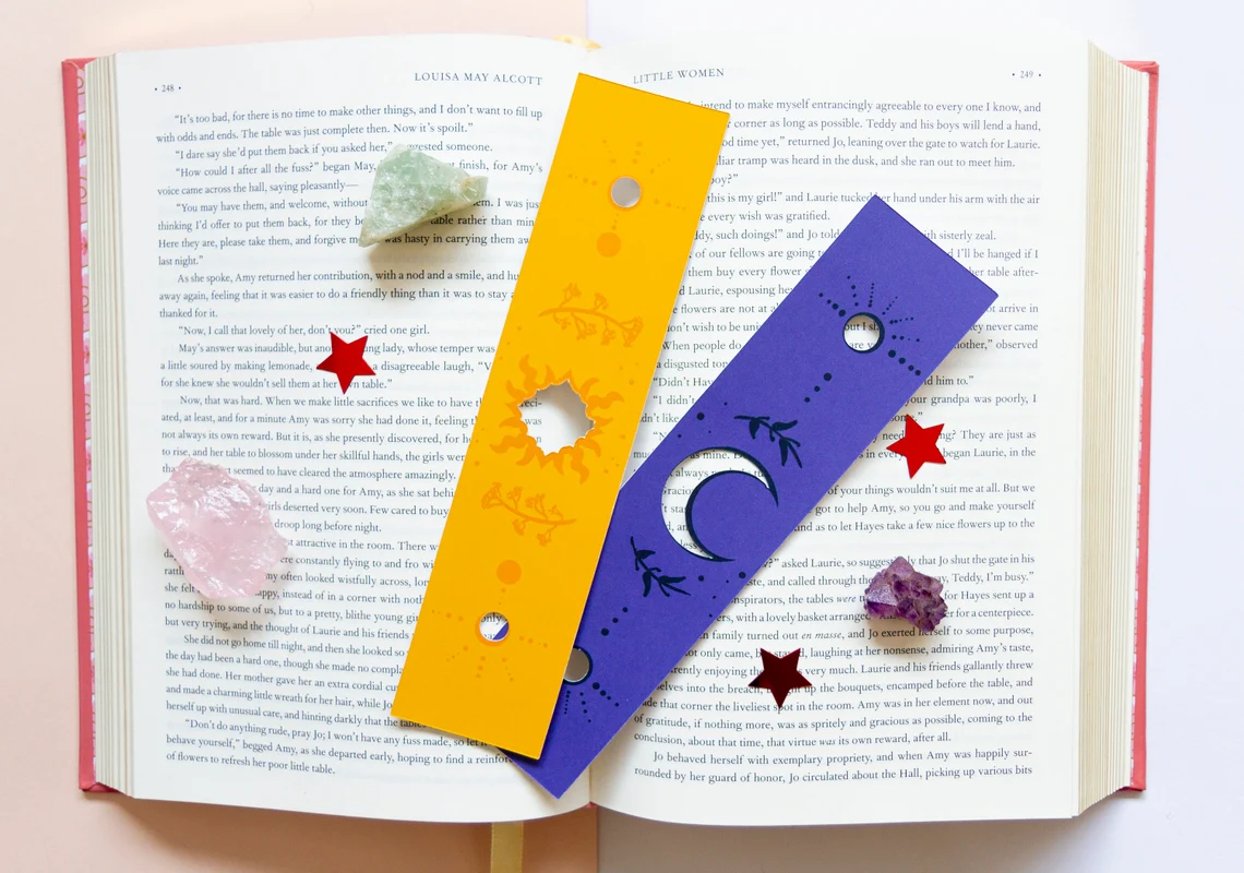 Image of an open book with a yellow bookmark with star cutouts and a dark purple bookmark with moon cutouts. 