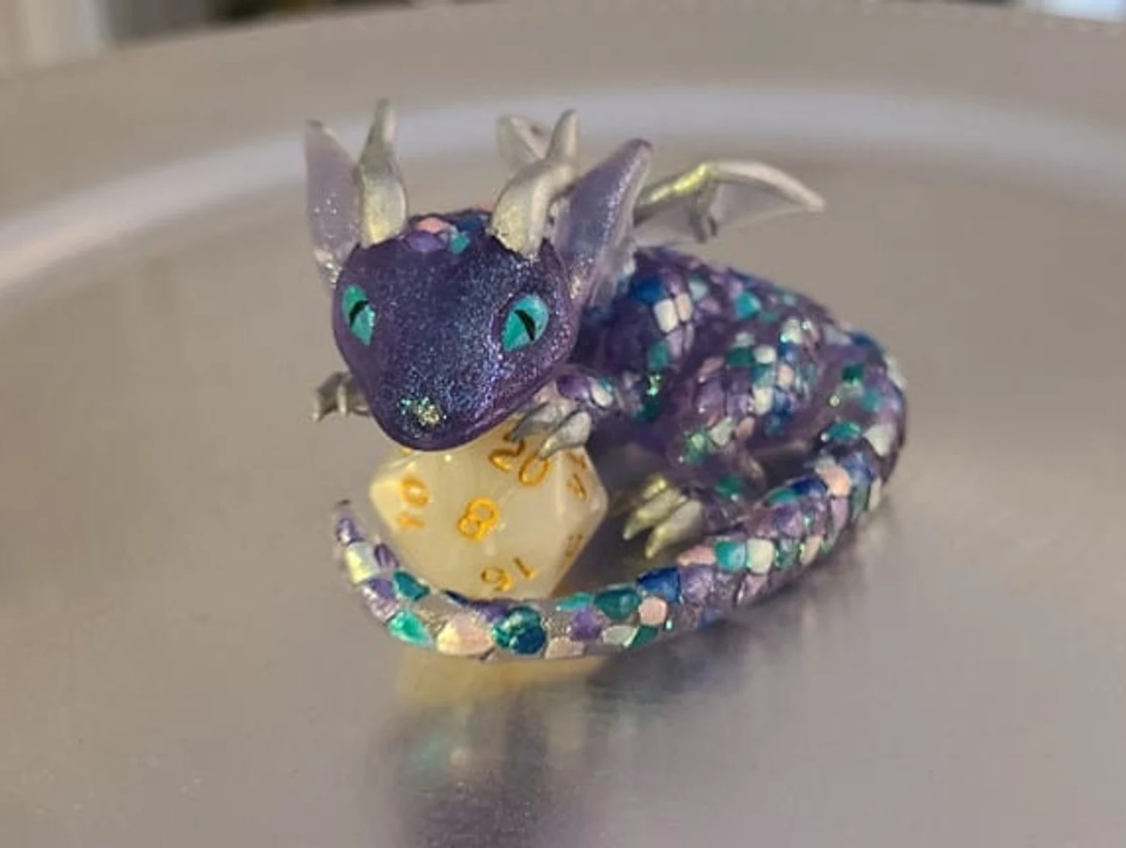 A photo of a small purple, blue, and silver dragon crouching over a yellow 20-sided dice. 