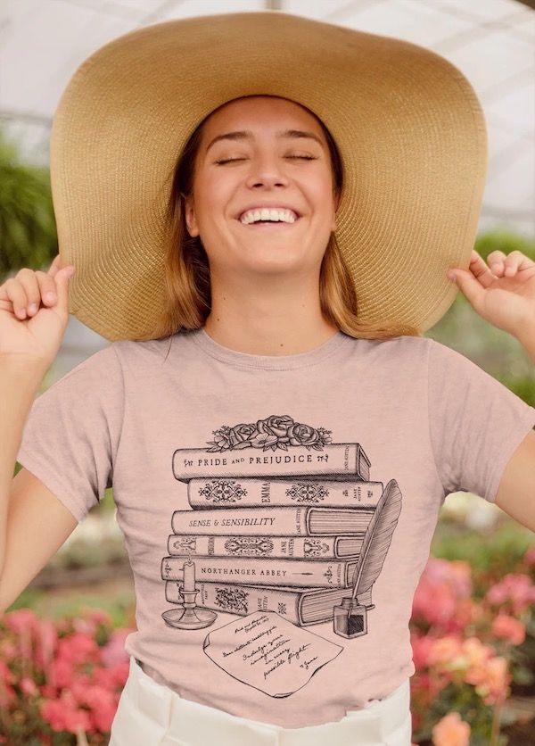 Photo of a woman wearing a hat and showcasing a light pink t=-shirt with printed a stack of books, a letter and a candle. The spines of the books read Pride And Prejudice, Emma, Sense And Sensibility, and Northanger Abby.