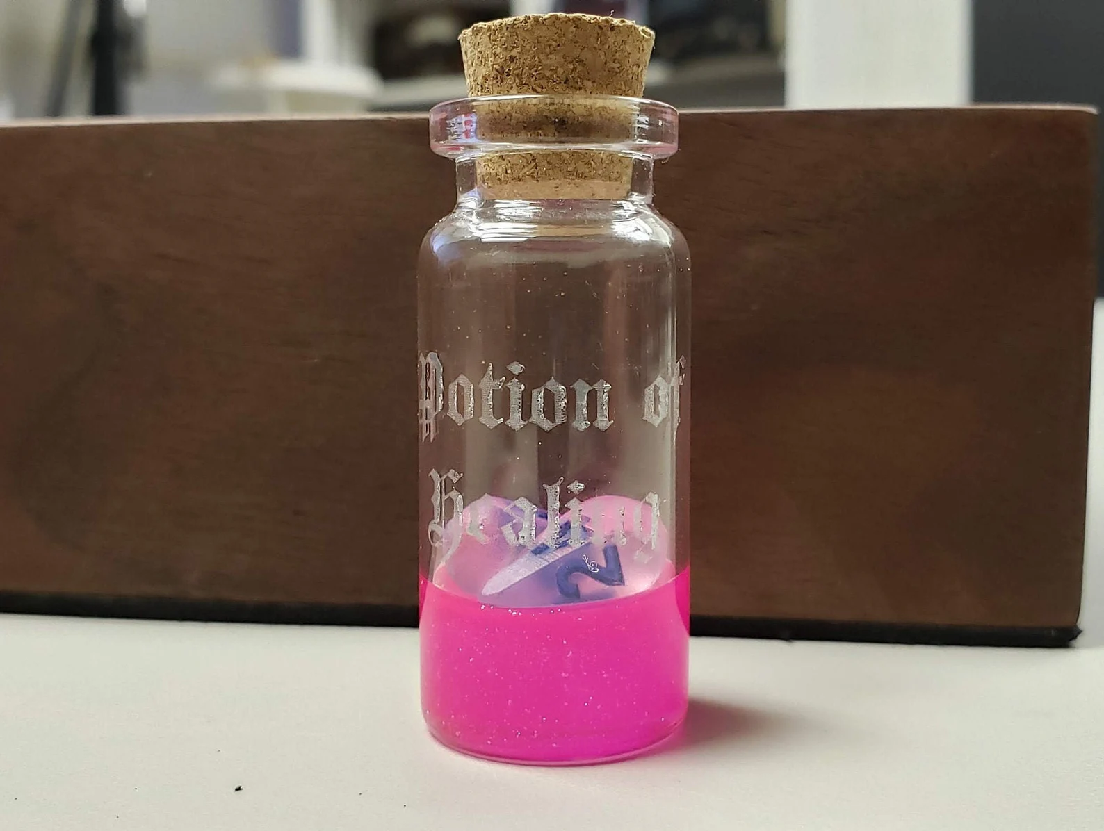 A photo of a small glass vial with a cork stopper and a pink, glittery band of color around the bottom. Potion of Healing is written on the glass vial. Two tear-shaped pink dice are inside the vial.