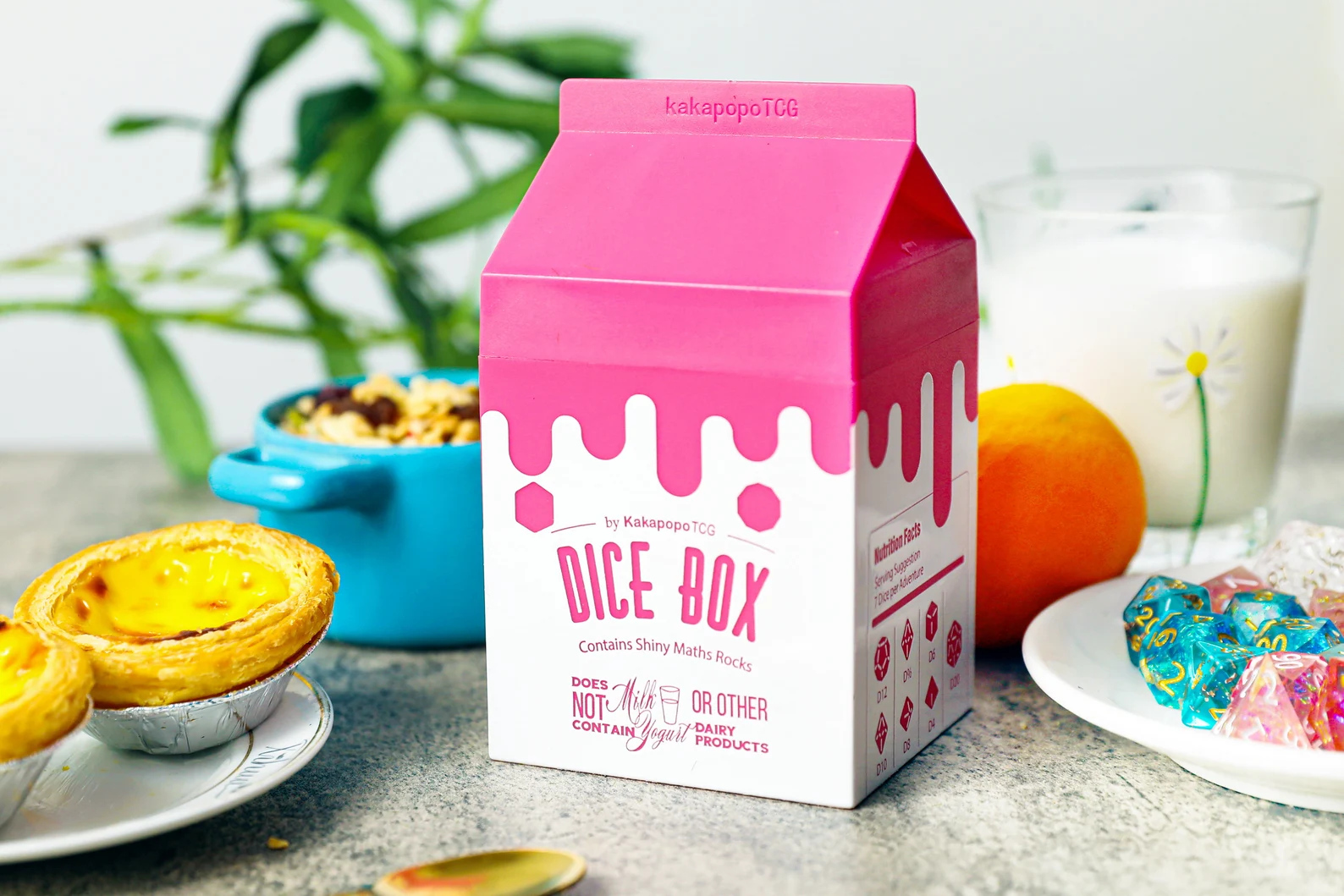 A photo of a plastic pink and white milk carton that says Dice Box on the front. Left to right the box is staged with egg tarts, granola, an orange, milk, and a plate of blue and pink dice. 