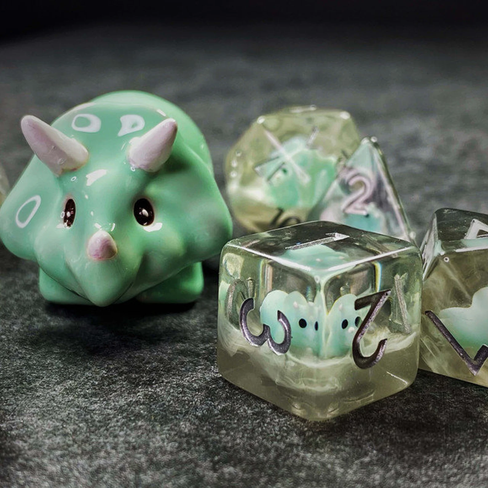 A photo of a set of four clear dice with silver numbers on the faces and teal Baby Triceratops inside. A larger ceramic teal Baby Triceratops is on the left of the photo.