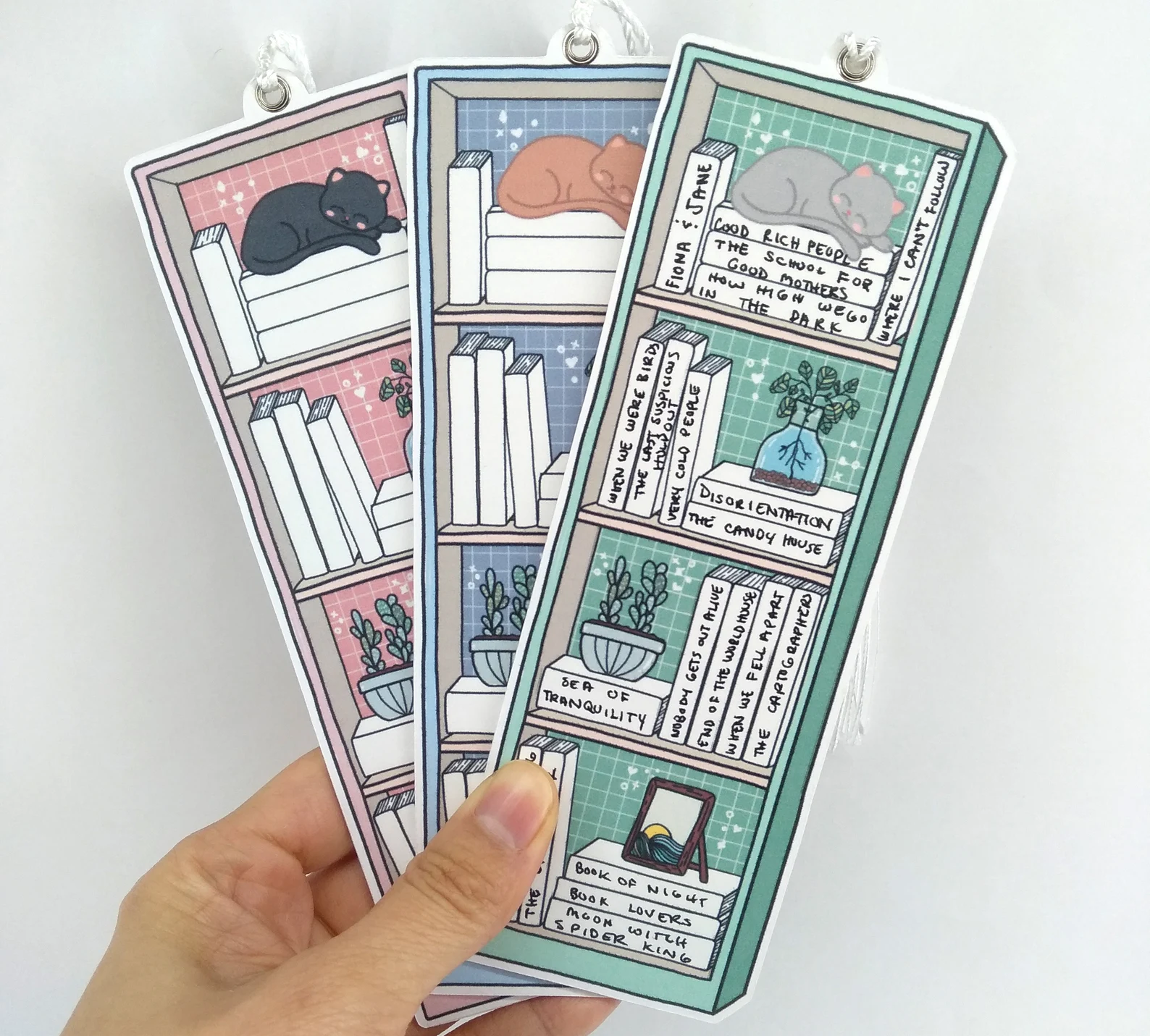 Three bookmarks depicting a bookshelf with books with blank spines and sleeping cats