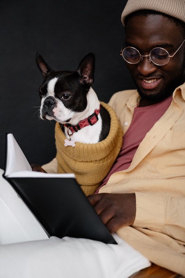 a black and white dog in a mustard knit sweater sitting on the lap of a person with black skin. The person is wearing round gold glasses and smiling, and they both appear to be reading a book together