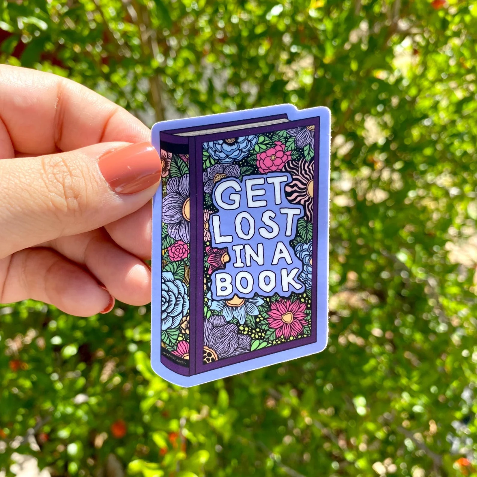 Image of a floral book sticker being held by a white hand. The text on the sticker says "get lost in a book."