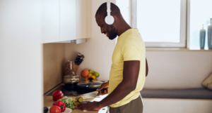 a photo of a Black man listening to headphones and smiling while cutting up vegetables in the kitchen