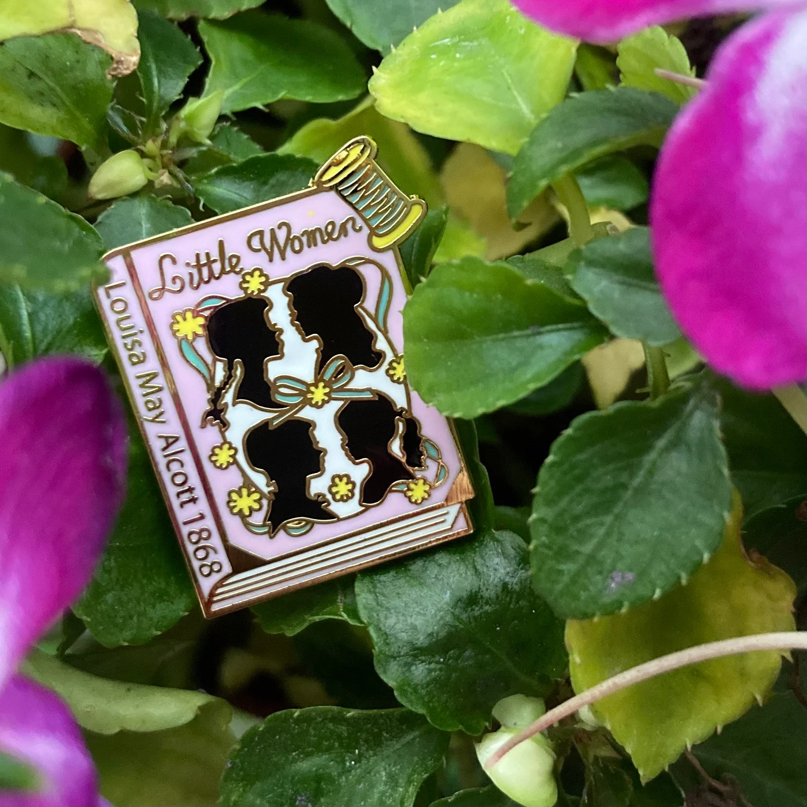 An enamel pin in the shape of a pink book with four profiles of women