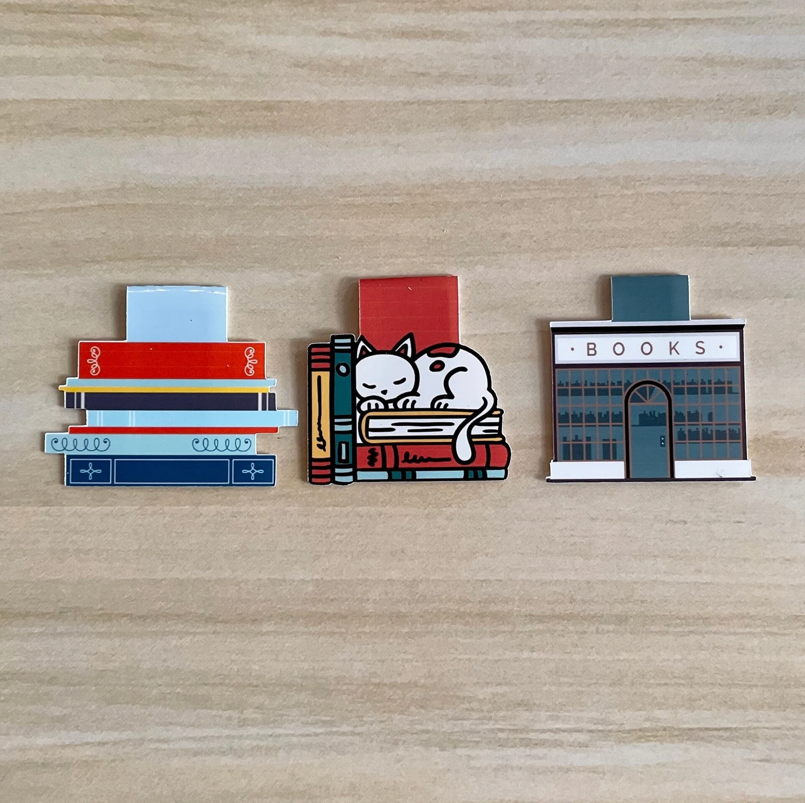 Image of three magnetic bookmarks. From left to right: a stack of books, a white cat on a stack of books, and finally, a bookstore. 