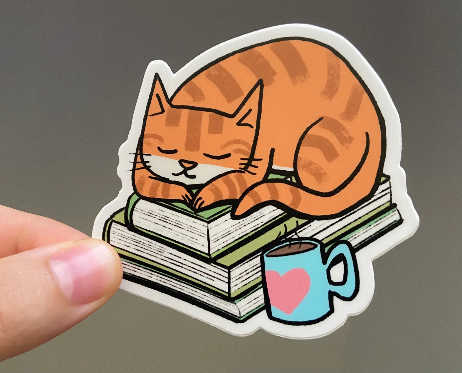 Image of a sticker which has an orange striped cat on a stack of books. 