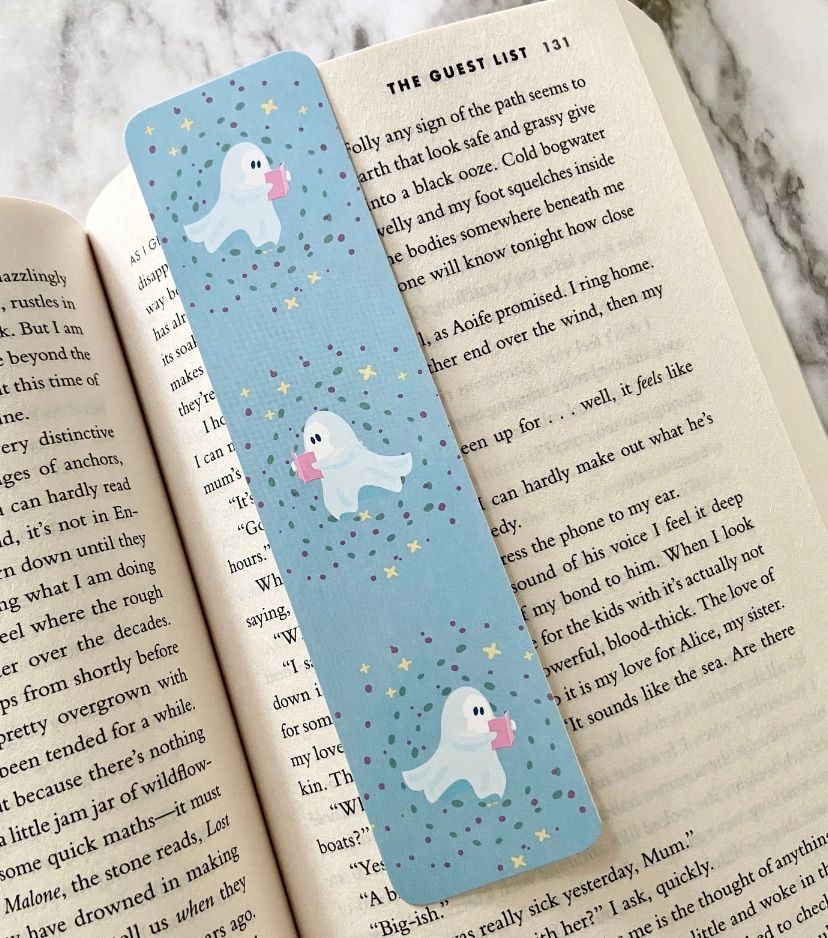 Image of a pastel bookmark inside an open book. There are images of ghosts reading on the bookmark. 