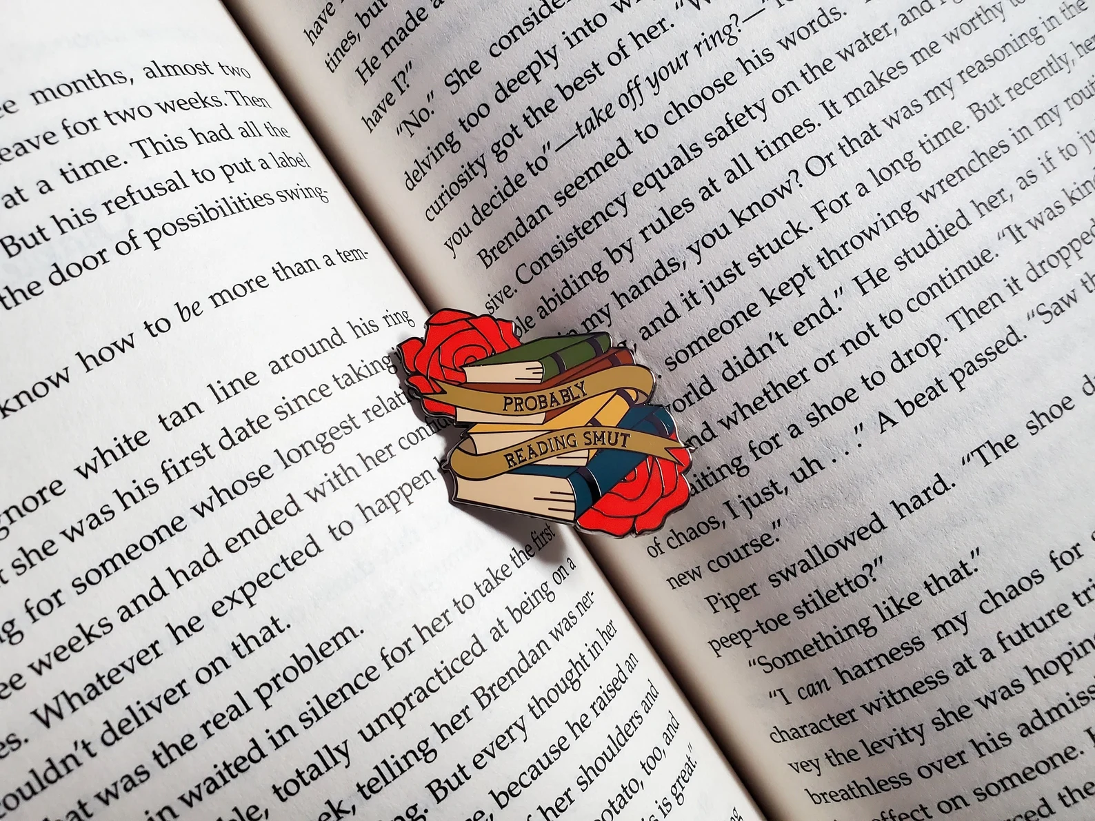 an enamel pin of a book stack and roses with the words "probably reading smut" as a banner