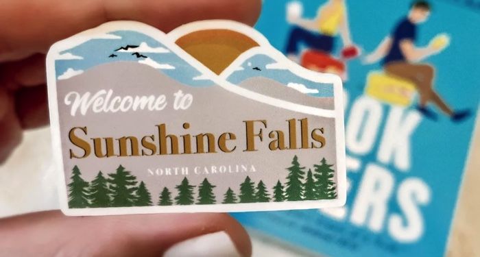 a sticker that says Welcome to Sunshine Falls with a paperback copy of Book Lovers by Emily Henry in the background