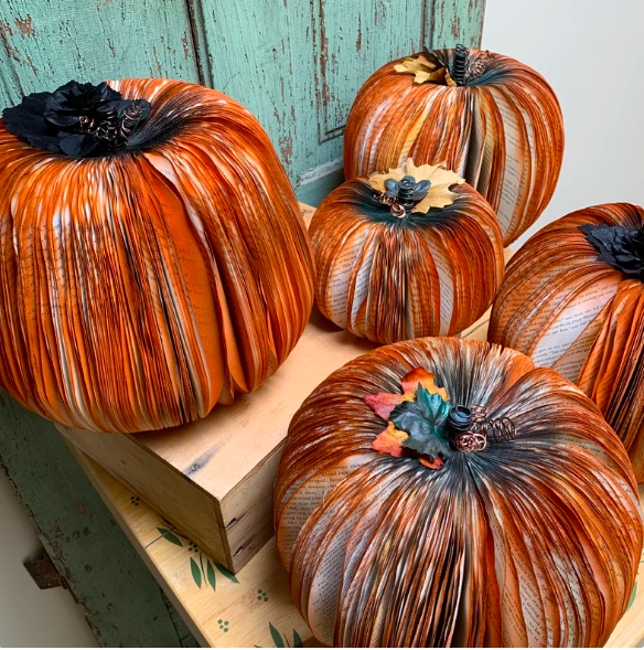 Five orange painted pumpkins topped with green leaves and flowers. The pumpkins are made from carved books.