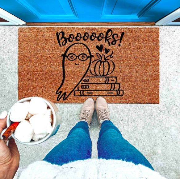 A brown doormat with a black outlined of a ghost in glasses next to a stack of books topped with a pumpkin and the text 