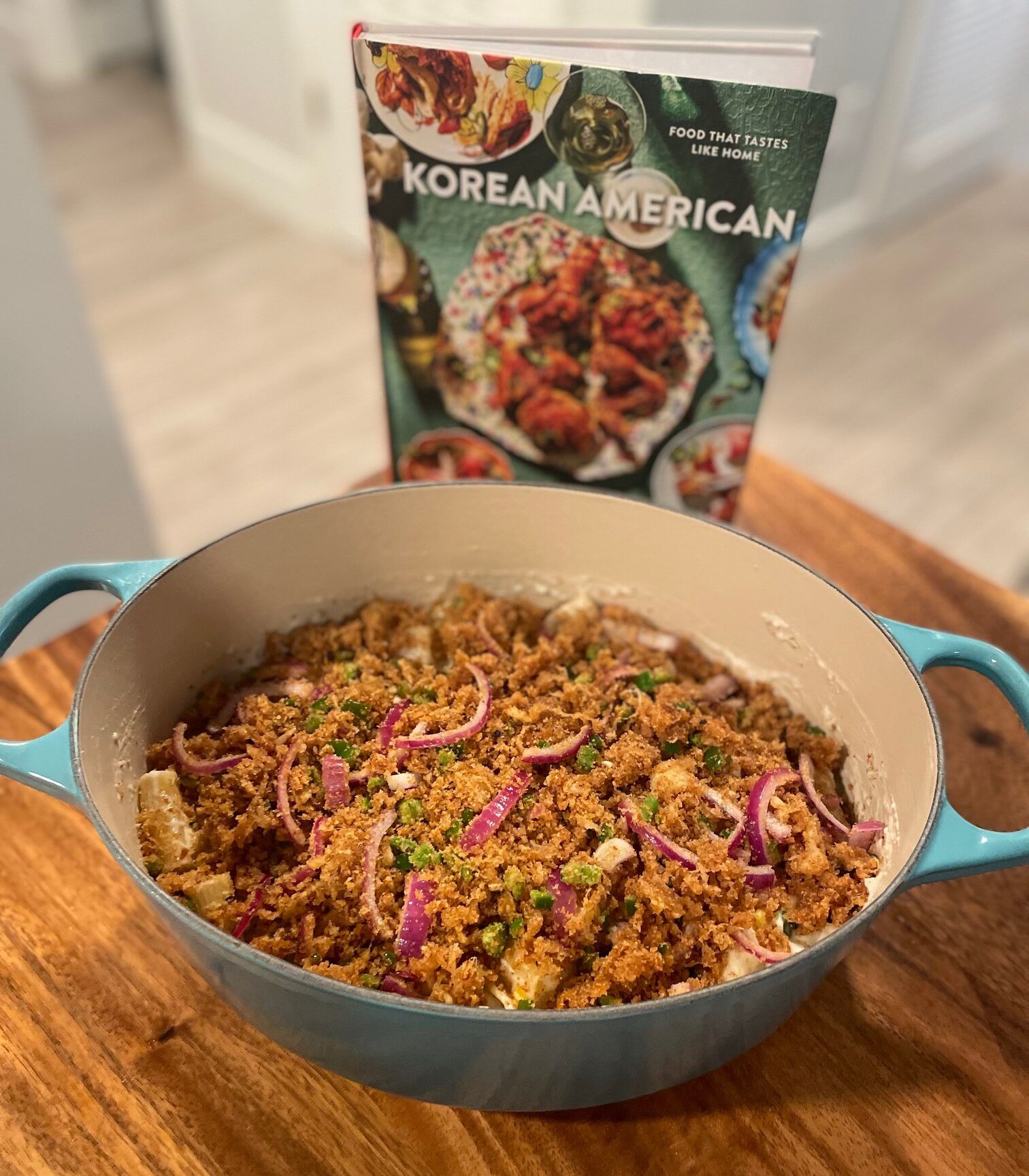 A teal dutch over full of a cheesy pasta topped with panko bread crumbs, red onion, and jalapeno. It sits on a wooden table in front of the cookbook Korean American