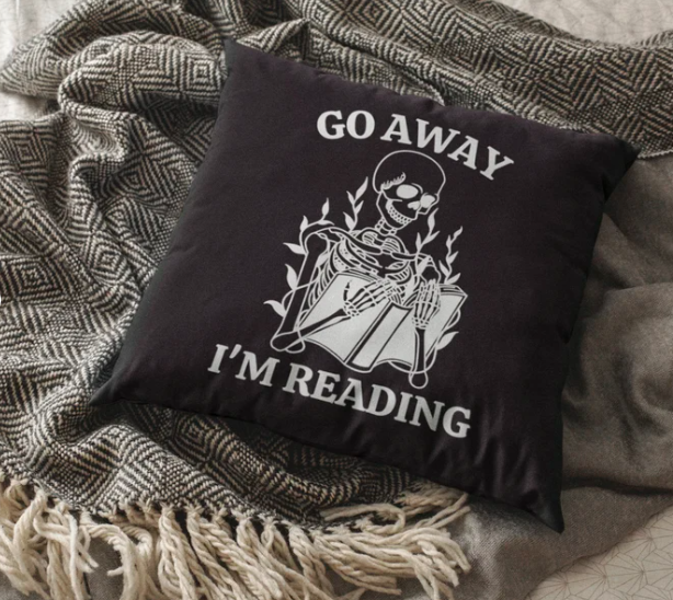 A black square throw pillow with an image of a skeleton reading and the text 