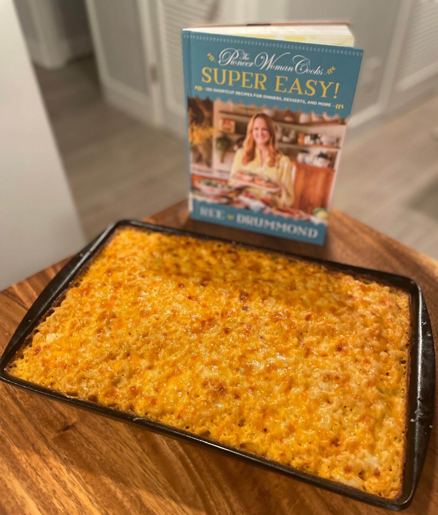 A sheet pan of mac and cheese sits on a wooden table in front of the cookbook The Pioneer Woman Cooks — Super Easy!