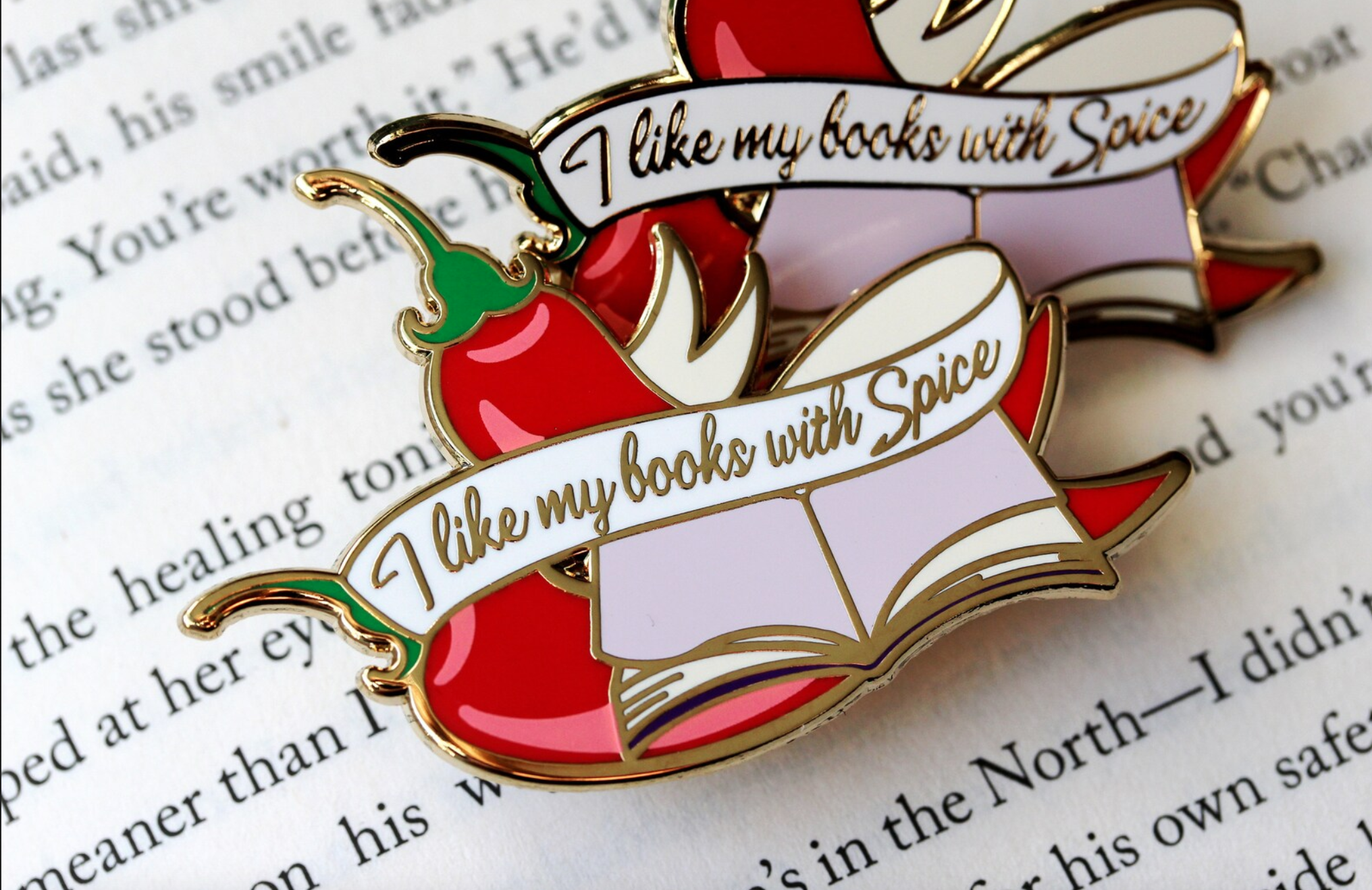 Enamel pin of an open book with chili peppers that says "I like my book with spice"