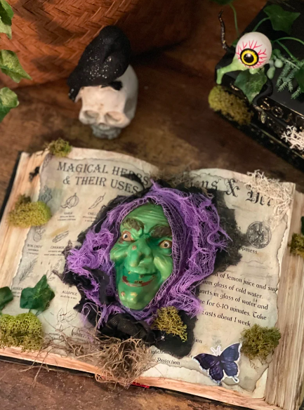 A book distressed to look spooky with the face of a witch poking through the center, green face and purple hair