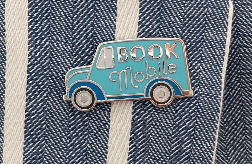 Image of a blue pin in the shape of a book mobile that says "book mobile" in silver. 
