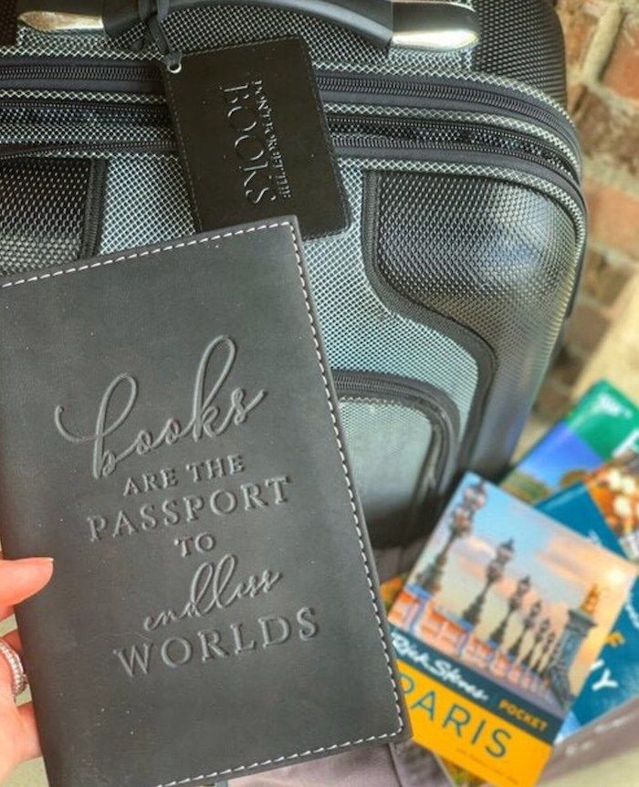 a hand holding a grey passport cover with embossed text that reads " Books are a passport to endless worlds." A grey piece of luggage with a matching luggage tag can be seen in the background
