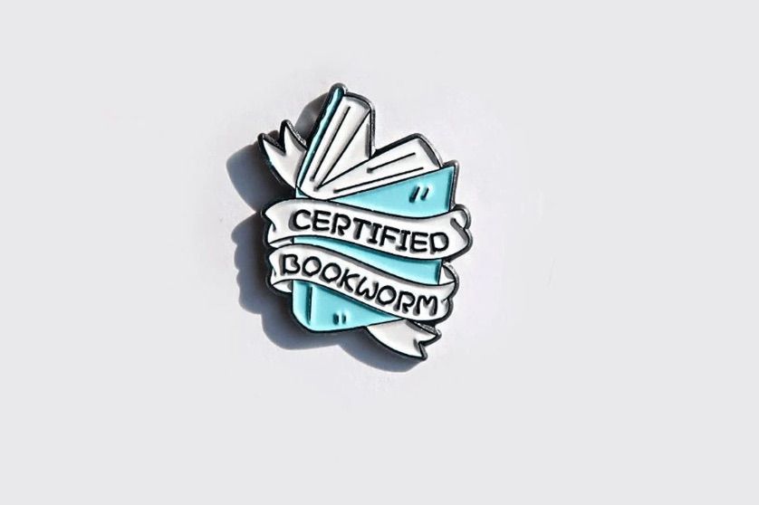 Image of a blue enamel pin that has a white banner reading "certified bookworm" on top of it. 